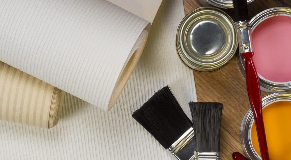 A close-up of two wallpaper rolls, three paintbrushes, and open cans of green, pink, and yellow paint on a wooden surface—perfect for anyone considering painting over wallpaper.