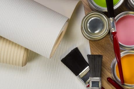 A close-up of two wallpaper rolls, three paintbrushes, and open cans of green, pink, and yellow paint on a wooden surface—perfect for anyone considering painting over wallpaper.