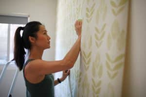 A woman applies wallpaper with a green leafy pattern to a wall in a bright room, holding a tool in her right hand—an alternative to painting over wallpaper for a fresh, vibrant look.